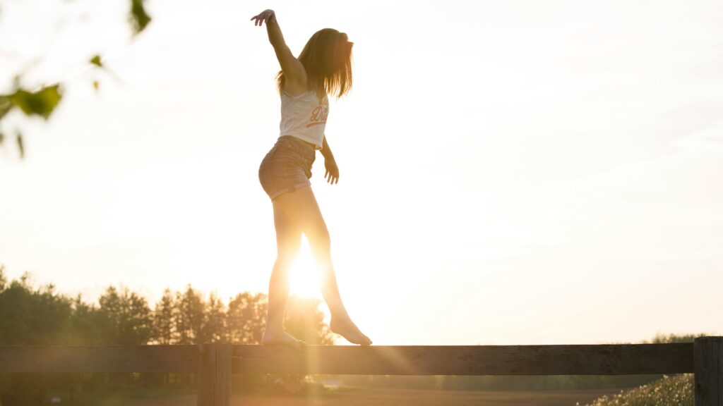 Person balancing barefoot along a wooden fence rail against a bright sunset