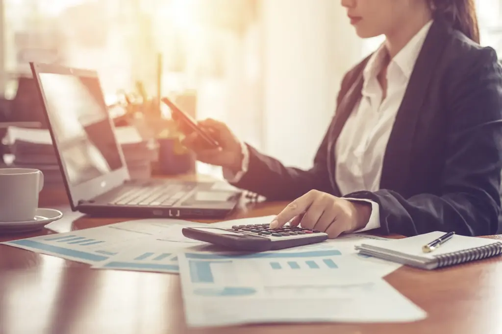 Financial adviser working with a calculator, laptop and printed charts while checking her phone