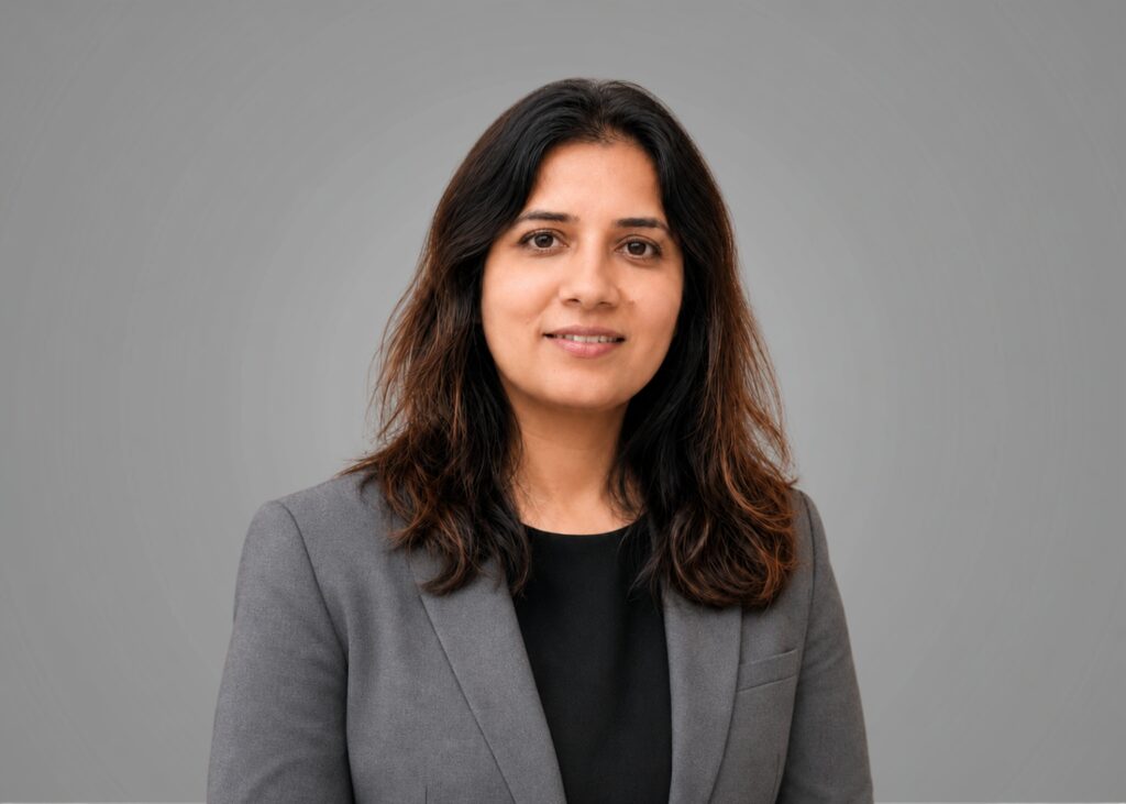 Headshot of a team member with long dark hair wearing a grey blazer over a black top against a grey background