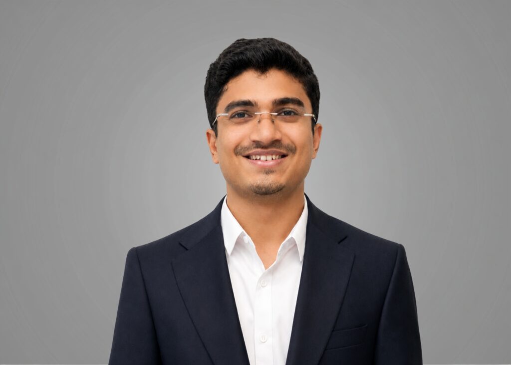 Headshot of a smiling team member in rimless glasses, navy suit and white shirt against a grey background