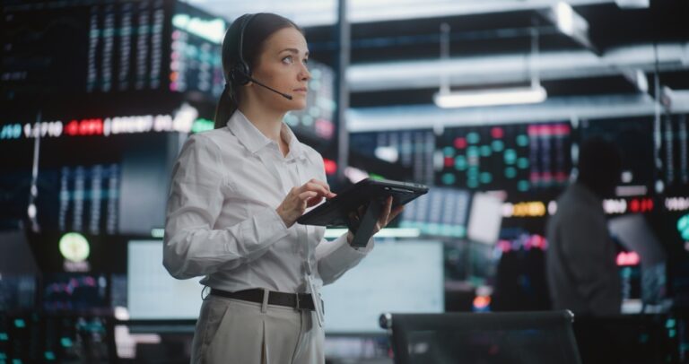 Trader with a headset and tablet on an exchange floor surrounded by market ticker screens