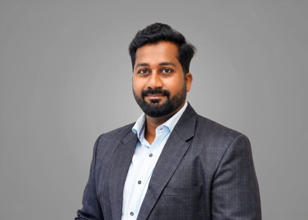 Headshot of a bearded team member in a checked charcoal suit and light blue shirt against a grey background
